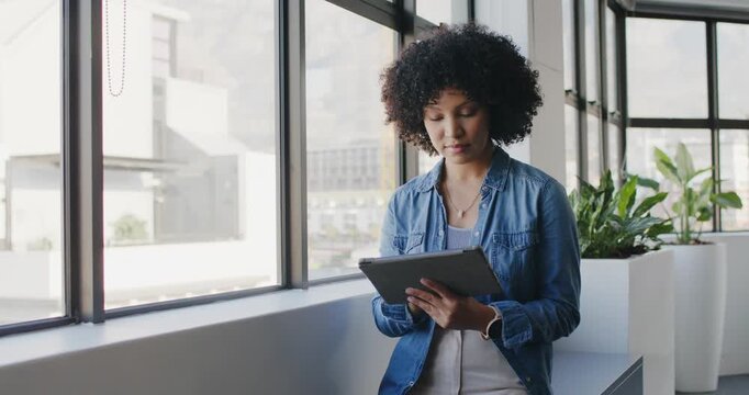 African American adult woman tapping tablet on alert, looking out office window while wearing denim