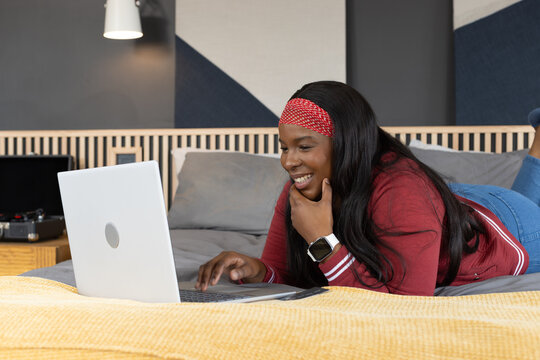 African American woman lying on stomach typing on silver laptop on bed wearing red jacket
