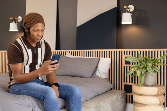 African American man sitting on edge of bed holding dark blue smartphone, wearing beanie