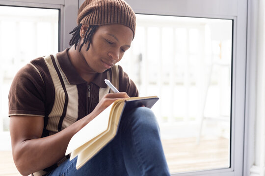 African American man sitting at home by glass door writing in journal with pen, wearing beanie