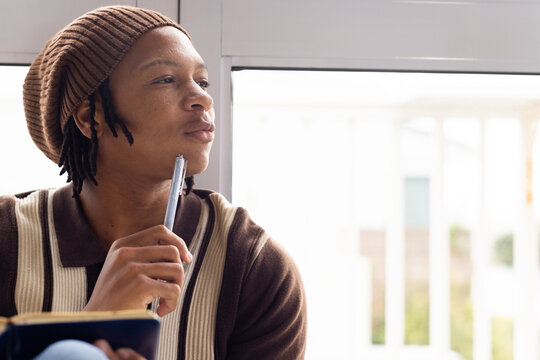 Male sitting by large window holding pen to chin, notebook, wearing striped sweater, copy space