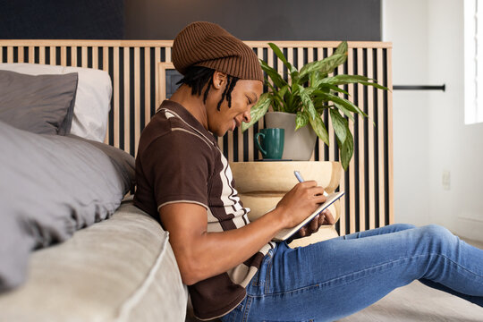 African American man sitting in bedroom wearing beanie striped polo and jeans writing in notebook