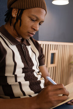 Man writing in cozy cafe nook wearing brown beanie and striped polo, holding notebook and pen