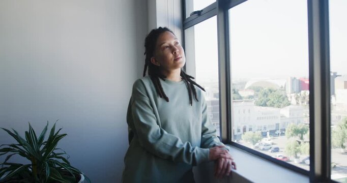 Nonbinary adult leaning on sill in green top, sunlight falling, turning to camera, smiling, plant