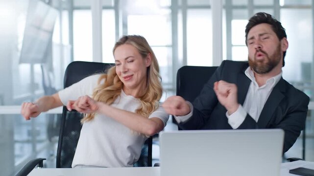 Two happy carefree business colleagues dancing same movements with hands listening music with laptop app having fun fooling enjoying joy with computer dance coworkers man with woman at office table
