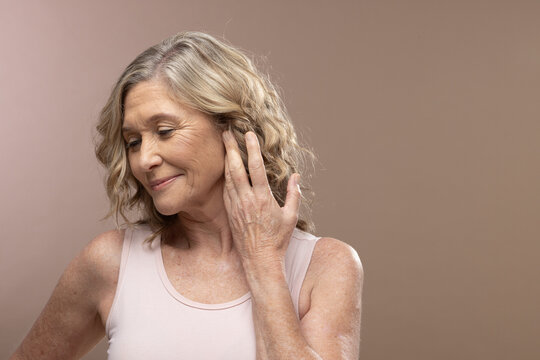Senior female posing at slight studio angle, touching hair, wearing pink tank top on beige backdrop