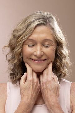 Mature woman pressing fingers to underside of chin while wearing sleeveless pale pink top in studio