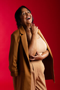 Senior African American woman laughing, wearing tan blazer and necklace in studio on red backdrop