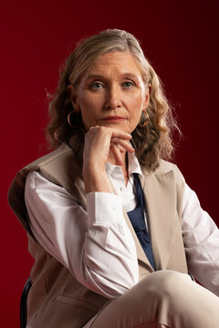 Senior woman posing in studio, resting chin on hand, wearing beige vest and gold hoops