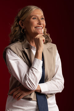 Senior woman posing in studio smiling upward, wearing white shirt beige vest navy scarf gold rings