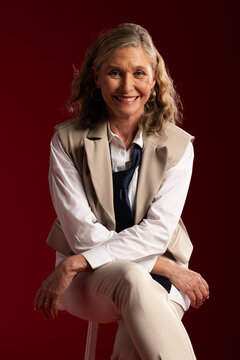 Senior woman sitting on stool wearing white shirt beige vest and navy necktie, smiling
