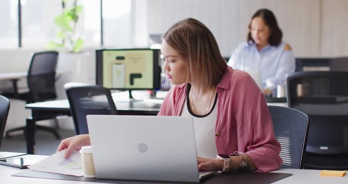 African American female coworkers while woman in pink top checking laptop comparing charts at desk