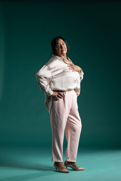 Senior woman posing in studio on teal backdrop, wearing satin blouse, slim pants, pumps, jewelry