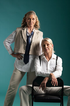 Senior female friends posing in studio wearing gender-neutral attire with black folding chair