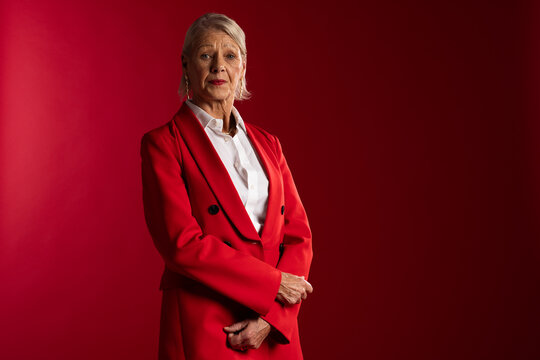 Senior woman posing in studio wearing red blazer white shirt drop earrings red backdrop, copy space