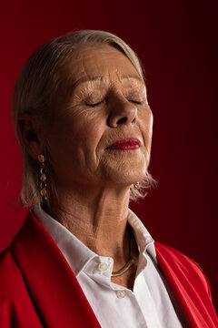 Senior woman closing eyes tilting head up in studio wearing red blazer gold jewelry red lipstick