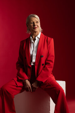 Senior woman sitting on white plinth in studio against red backdrop wearing red suit, earrings