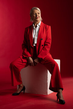 Senior woman sitting on white block in studio, wearing red suit, black pumps, showing ring