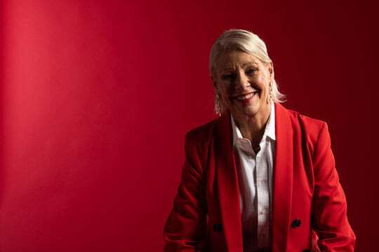 Senior woman sitting on right in studio smiling wearing red blazer and drop earrings, copy space