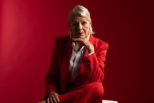 Senior woman sitting on white cube in studio wearing red tailored suit, gold earrings, red backdrop