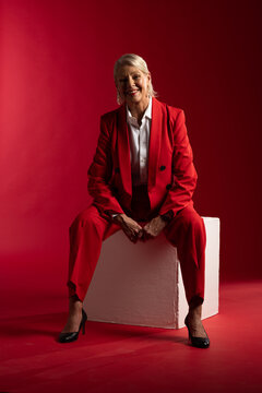 Senior woman sitting on white block in studio against seamless red backdrop, wearing red suit