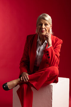 Senior woman posing thoughtfully on white pedestal in studio wearing red suit and hoop earrings