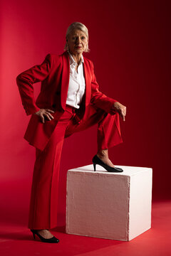 Senior woman posing in studio wearing red suit and white blouse, resting foot on white cube