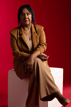Senior African American woman sitting on white cube in studio wearing tan suit and chain necklace
