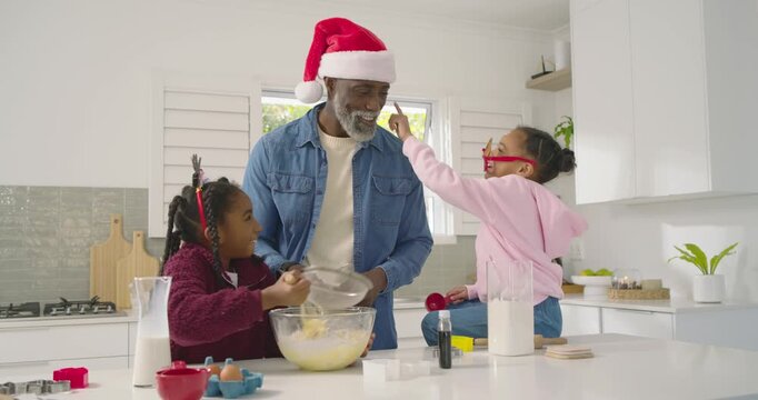African American man with two youth girls pointing, guiding, girls whisking batter at island, sieve