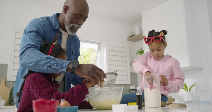 African family dad placing bowl, guiding youth girls sifting and making batter at kitchen island