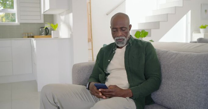 Senior African man sitting on gray sofa holding blue smartphone, tapping and smiling at content