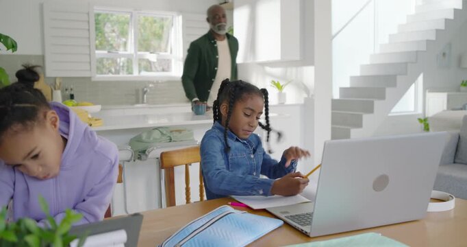 Senior man seeing homework guiding girls at kitchen table pointing to laptop as denim girl writing
