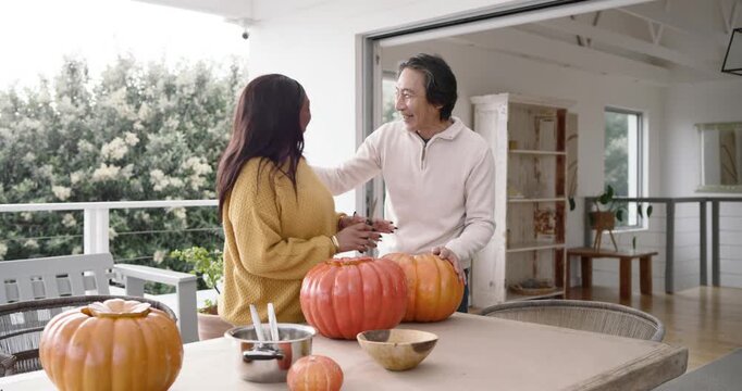 Senior Asian man carrying pumpkin in, woman greeting, carving pumpkins with knife at patio table