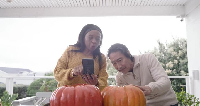 Diverse couple showing phone, senior Asian man and woman moving pumpkins on porch for fall