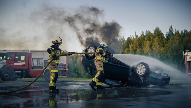 Car Crash: Two Brave Firefighters in Protective Gear Spray a Fire Hose Onto a Burning Overturned Car at a Road Accident Scene, Thick Black Smoke Rising as the Fire Engine Stands by for Rescue.