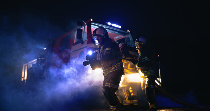 Team of Male Firefighters in Helmets and Reflective Uniforms Drag a Hose Through Smoke During a Night Car Crash Rescue Operation, With a Fire Truck and Blue Emergency Lights Behind.