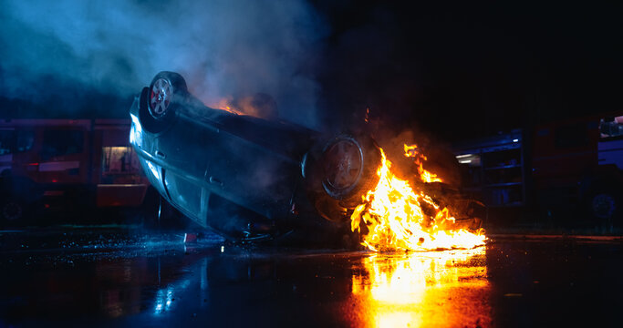 Overturned Car Engulfed in Flames at Night After a Deadly Traffic Collision, With Fire Engines Arriving on the Emergency Scene as Rescue Crews Respond to the Vehicle Rollover Accident.