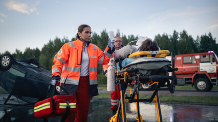 Obraz premium Paramedic Team Transporting Injured Victim on Stretcher With Iv Drip and Oxygen After Car Crash, Overturned Vehicle and Fire Truck at Traffic Accident Scene During Evening Rescue Operation