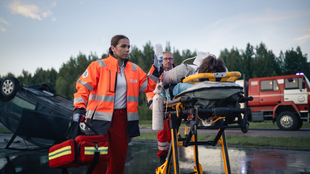 Paramedic Team Transporting Injured Victim on Stretcher With Iv Drip and Oxygen After Car Crash, Overturned Vehicle and Fire Truck at Traffic Accident Scene During Evening Rescue Operation
