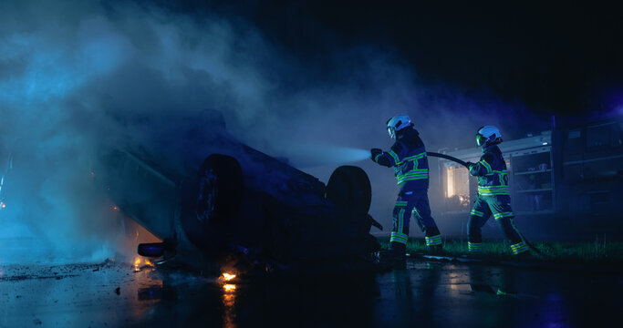 Two Firefighters Direct Water Hose at Smoking Overturned Vehicle After Car Crash at Night, Fire Truck Behind Rescue Workers Extinguishing Flames at Traffic Accident Scene on Road