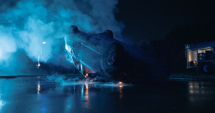 Overturned Car Smolders on the Road After a Severe Traffic Accident at Night, Smoke and Flames Rise From the Wrecked Vehicle as a Fire Truck Stands by at the Car Crash Scene Awaiting Rescue Workers