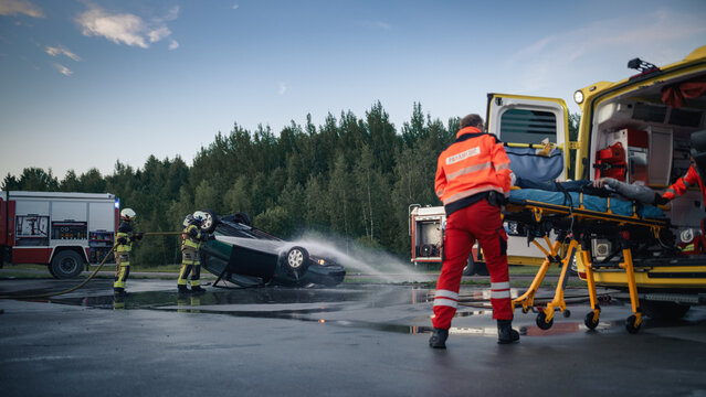 Wide View of Car Crash Rescue Scene: Paramedic Team Lifts Injured Victim on Stretcher Into Ambulance While Two Firefighters Hose Down Overturned Vehicle, Traffic Accident Response at Dusk