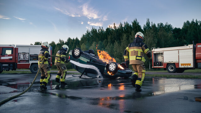 Three Firefighters Rush to Extinguish a Burning Car After a Traffic Accident, Rescue Workers Spray Water on the Upside Down Vehicle Engulfed in Flames at a Car Crash Site Between Two Fire Trucks