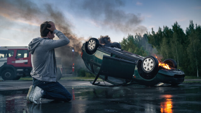 Shocked Young Man Kneels on the Road Holding His Head While Watching a Burning Upturned Vehicle After a Car Crash, Traffic Accident Survivor Waits for Firefighters and Rescue Workers