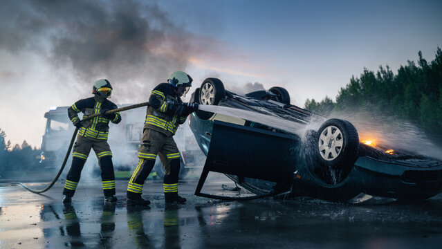 Two Firefighters Spray Water From Hose to Extinguish Flames on Overturned Burning Car After Night Traffic Accident, Rescue Crew Battling Fire at Car Crash Scene With Emergency Fire Truck in Background
