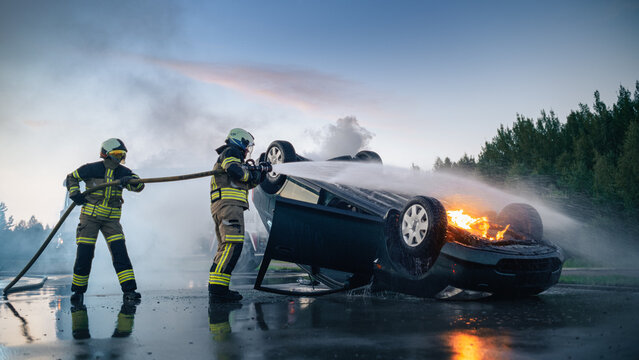 Firefighter Team Extinguishing Burning Rolled Over Vehicle With Fire Hose After Serious Car Crash at Dusk, Rescue Workers Putting Out Flames and Smoke at Traffic Accident Scene on Road