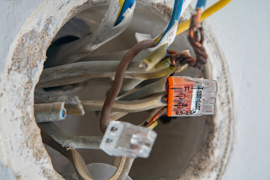 Macro shot of an open flush-mounted round electrical junction box in a wall filled with stripped cables, colorful wire insulation, copper conductors, and transparent push-in wire connectors.   