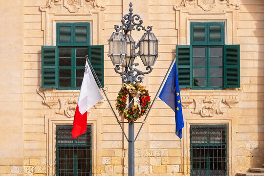 Vintage street lamp decorated with festive Christmas wreath, national flag of Malta and European Union flag against historic building facade.