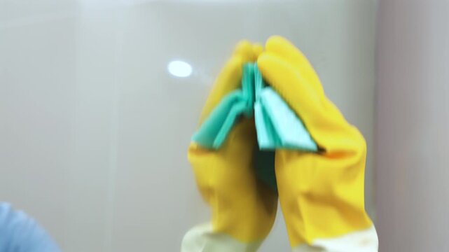 Close-up of housekeeper hand wearing yellow rubber glove wiping mirror with blue cloth. Glass surface cleaning, house maintenance, domestic chores, and hygiene concept.