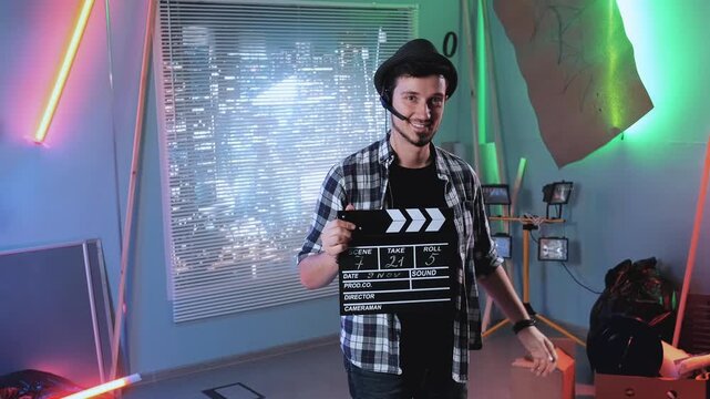 A man in a plaid shirt and black headset holds a movie clapper board while posing indoors in a professional studio with colorful lighting and cityscape at night.